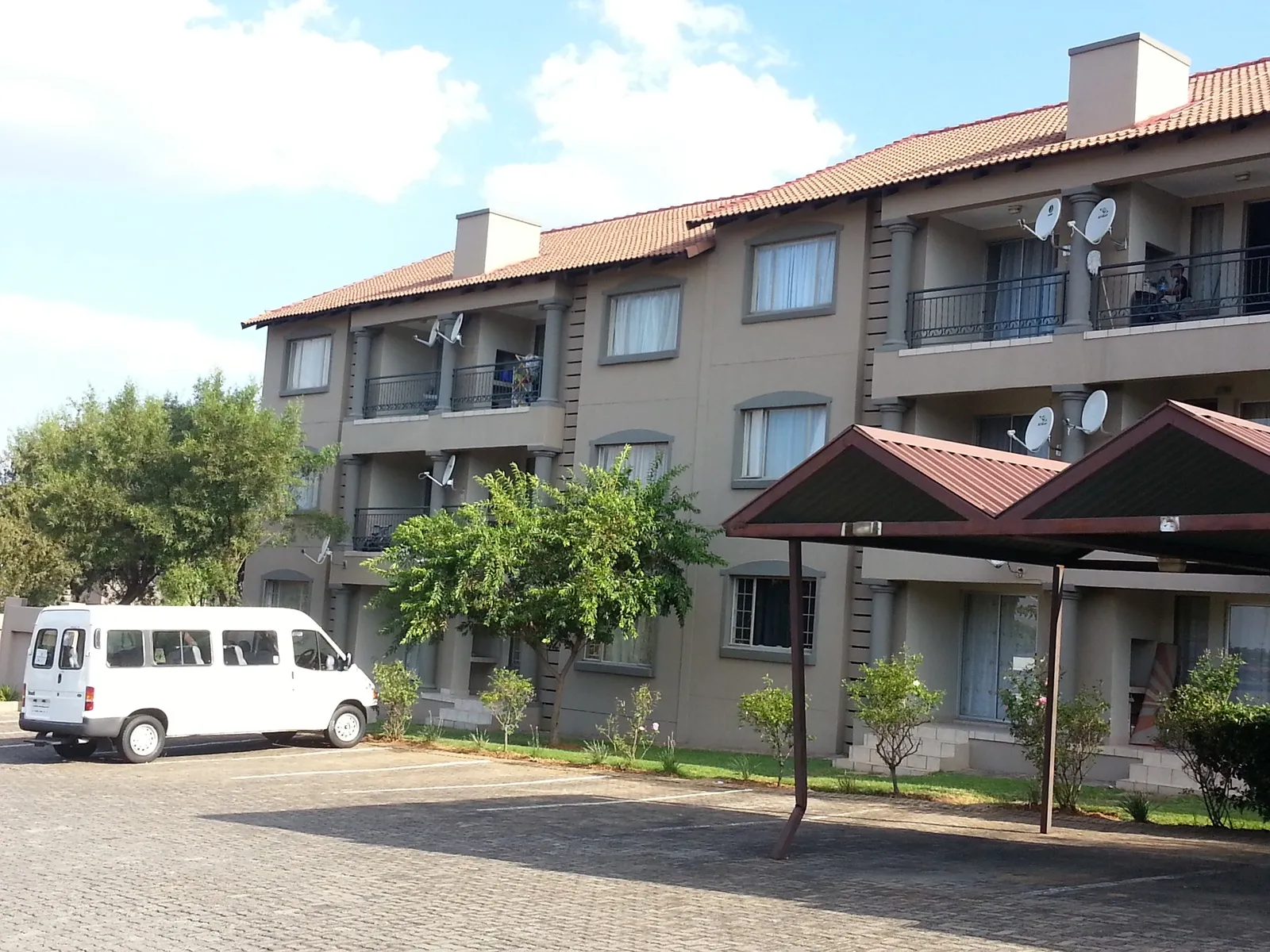 A beige three-storey townhouse complex with tiled roof and balconies, covered carports and a paved parking area